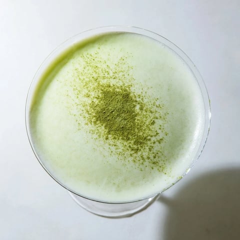 A close-up of a vibrant Matcha Martini cocktail in a chilled glass, garnished with matcha powder and a glossy white chocolate shave, its creamy pale green surface frothy and inviting.
